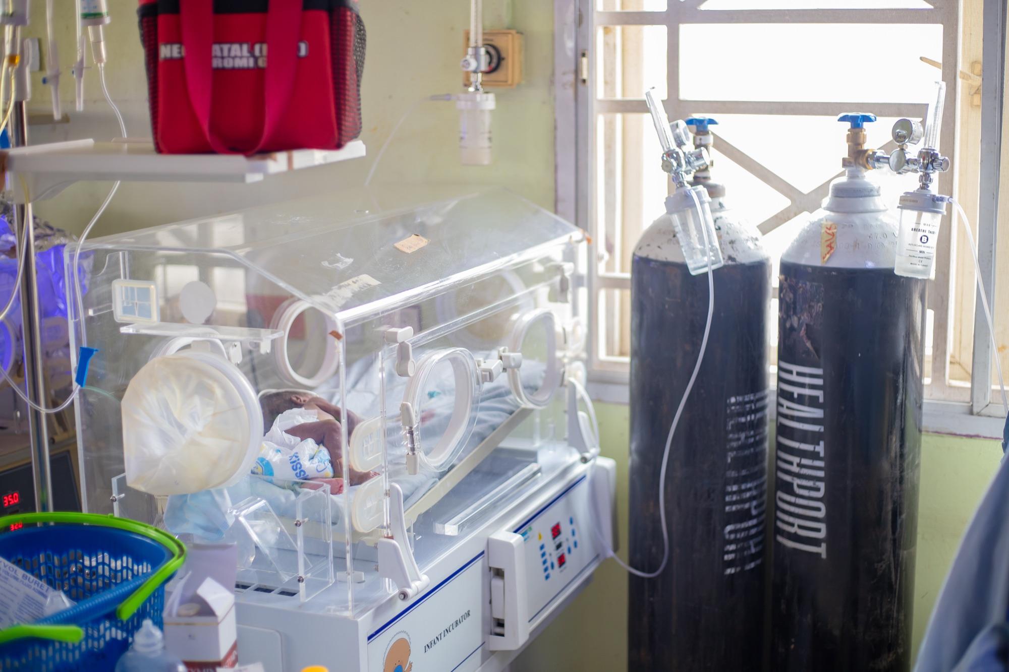 A newborn in a NICU incubator beside HealthPort-branded oxygen cylinders, infrastructure and care in one frame.